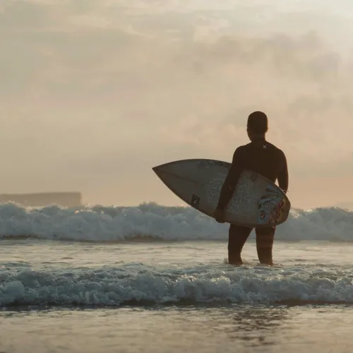 Sober Companion & Coach Taylor Wilson, standing on the beach holding a surfboard